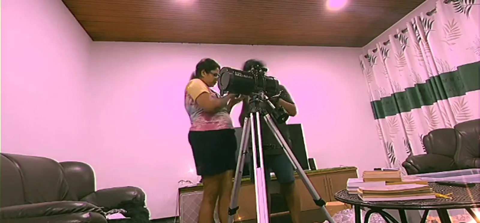 Setting up a large reflector telescope on a tripod in a sitting room lit pink, aligning the finder by hand.