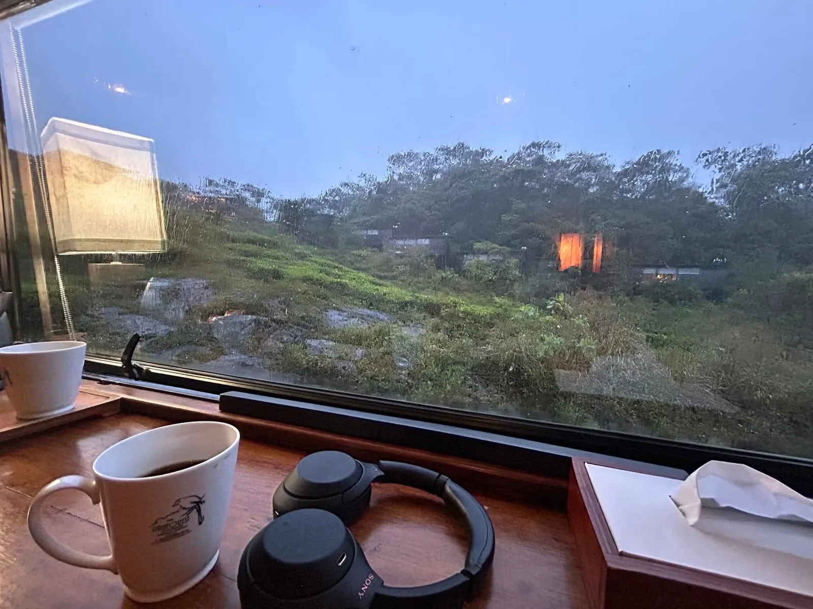 A mug of black coffee and over-ear headphones on a wooden sill, looking through rain-flecked glass at a green hillside at dusk.